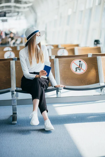 Itsevarma liikenainen naamiossa istuu lentokentällä, jolla on passi ja lentoliput, pitää etäisyyttä pandemiaan. Odotan lentokentällä. Matkakonsepti Nainen katselee lippuja — kuvapankkivalokuva