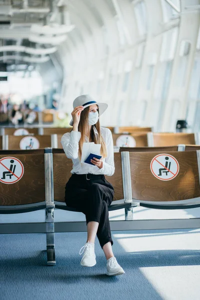 Nuori nainen turisti yllään kasvonaamio tilalla passi ja lippu ja käyttämällä älypuhelin, istuu lentokentällä tai rautatieasemalla tyhjällä merkitty tuolit uuden normaalin ja sosiaalisen covid etäisyyden sääntelyn — kuvapankkivalokuva
