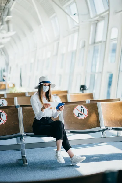 Ung jente, forretningskvinne står ved vinduet på en moderne flyplass med billetter og telefon, og ser inn i kameraet. Portrett av en vakker jente i maske. Forretningsreise med fly – stockfoto