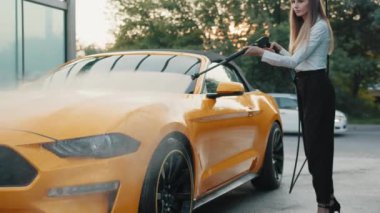 Başarılı bir iş kadını beyaz gömlek ve siyah pantolon giyer, sarı arabasını açık hava yıkama servisinde yüksek basınçlı hortumla elle yıkar. Araba yıkama konsepti. Araba Servisi