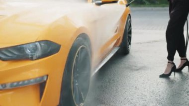 Kafkasyalı bir kadının, yüksek basınçlı suyla, açık hava oto yıkama servisinde, sarı lüks arabasını yıkarken çekilmiş yatay görüntüsü.