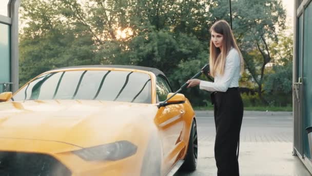 Femme d'affaires joyeuse portant sur une chemise blanche et un pantalon noir laver sa voiture jaune avec un pistolet à eau sur la station de lavage libre-service de voiture à l'extérieur, rinçant la saleté. Concept de lavage de voiture — Video