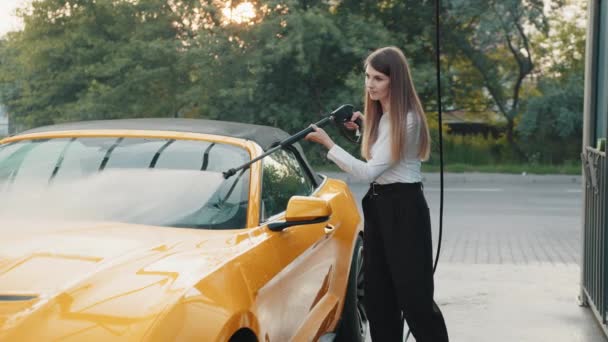 Manuelle Autowäsche. Junge Frau in Business-Outfit, Hemd und Hose wäscht ihr luxuriöses gelbes Fahrzeug mit Hochdruckwasserpumpe bei der Autoreinigung im Freien. Konzept Autowäsche. — Stockvideo