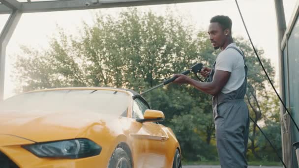 Bello africano giovane in tuta protettiva, lavando la sua auto gialla manualmente con acqua tubo ad alta pressione a servizio di self-wash all'aperto. Concetto di lavaggio auto. L'acqua è stata diffusa alla macchina — Video Stock