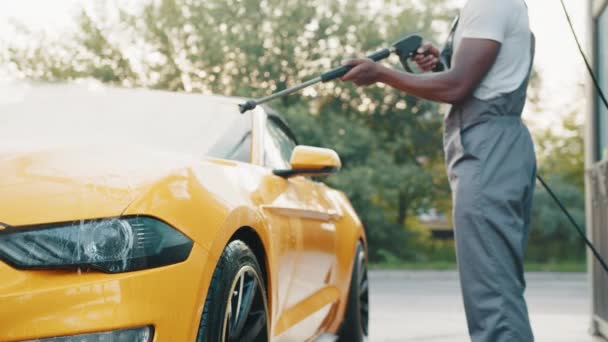 Autowaschanlage im Freien. Afrikanischer Typ in grauen Overalls und weißem T-Shirt, der sein gelbes Luxusauto mit Wasserpistole auf einer Autowaschanlage unter freiem Himmel wäscht. Autowäsche-Konzept, Car Service — Stockvideo