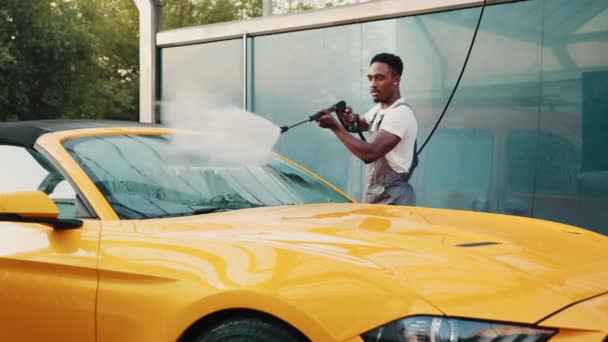 Ручная автомойка. Красивый африканский молодой человек моет свой роскошный желтый автомобиль с водяным насосом высокого давления на автомобильной уборке самообслуживания на открытом воздухе. Концепция мойки автомобилей, сервис — стоковое видео