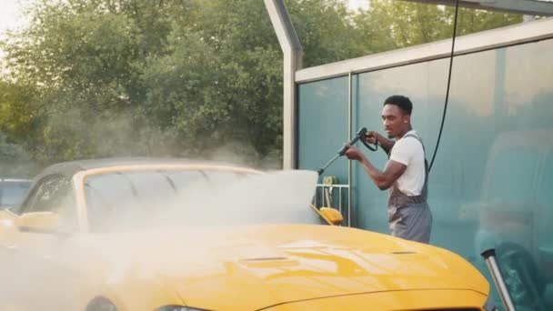 Conceito de lavagem de carros, Serviço. Lavagem manual de carro. Bonito jovem africano lavando seu luxuoso veículo amarelo com bomba de água de alta pressão na limpeza de automóveis self service ao ar livre — Vídeo de Stock