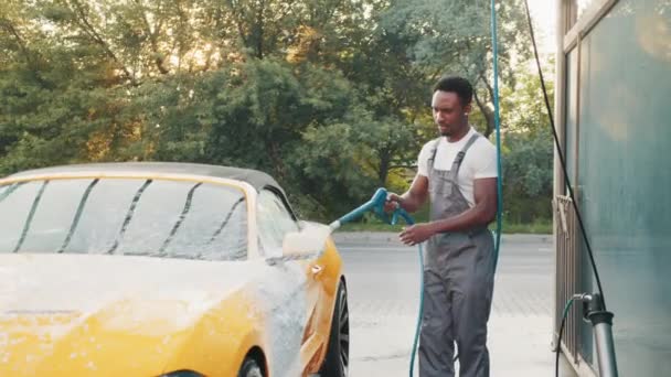 Egy jóképű, szakállas afrikai fiatalember portréja, aki habszivaccsal mossa a sárga autóját az önkiszolgáló autómosóban a szabadban. Tiszta autó koncepció. Autómosó. Autómosás koncepció, szolgáltatás — Stock videók