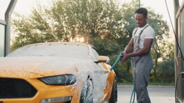 Dışarıda oto yıkamada sarı arabasını köpükle yıkayan yakışıklı sakallı genç Afrikalı adamın portresi. Temiz araba konsepti. Araba yıkama. Araba yıkama konsepti, servis. Manuel oto yıkama.
