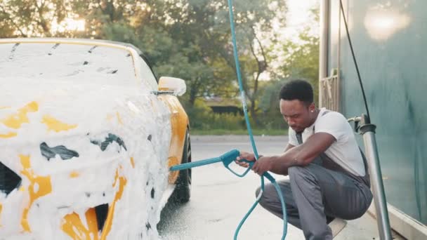 Τροχός αυτοκινήτου, χείλος ή κράμα, πλύνετε σε εξωτερικούς χώρους. Καθαρισμός αυτοκινήτου με τζετ νερού. Οριζόντια βολή του ευχάριστου Αφρικανού με λευκό t-shirt και φόρμες, πλύσιμο του κίτρινου τροχού του αυτοκινήτου με νερό υψηλής πίεσης jet — Αρχείο Βίντεο