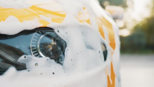 Close-up horizontaal schot van gele auto koplamp met reinigingsschuim, gewassen bij carwash service buiten met hogedruksproeier. Schone auto concept. Auto wassen buiten. Schuim na het wrijven van de auto — Stockvideo