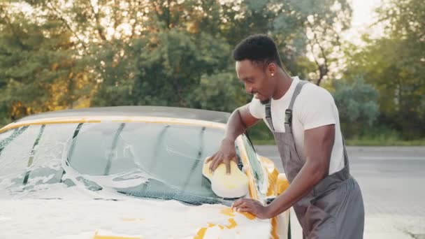 Jeune homme afro-américain attrayant laver sa voiture de luxe jaune dans une station de lavage de voiture libre-service à l'extérieur avec mousse de nettoyage et éponge jaune. Concept de voiture propre. Moussant après avoir frotté la voiture. — Video