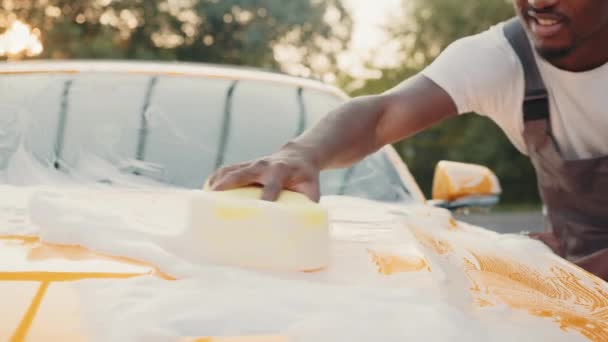 Closeup image of hand of young African man with yellow sponge washing headlight of his car at a self-serve car wash outdoors. Car covered by foam. Car wash outdoor. Foaming after rubbing the car. — Stock Video