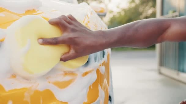 Primer plano imagen recortada de la mano del joven africano con faro de lavado de esponja amarilla de su coche en un auto-servicio de lavado de coches al aire libre. Coche de lujo cubierto de espuma. Espuma después de frotar el coche. — Vídeos de Stock