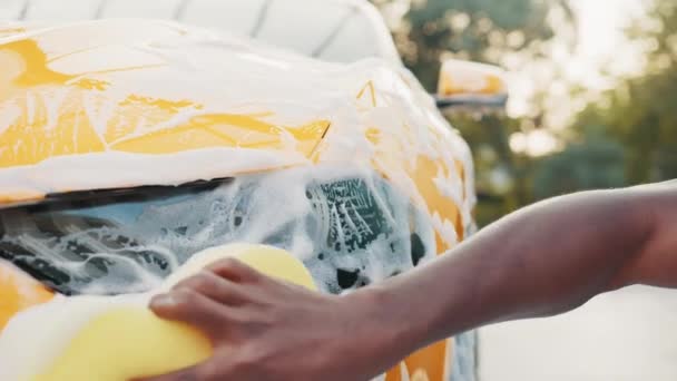 Gros plan recadré image de la main du jeune homme africain avec éponge jaune lavant phare de sa voiture à un lavage de voiture libre-service à l'extérieur. Voiture de luxe recouverte de mousse — Video