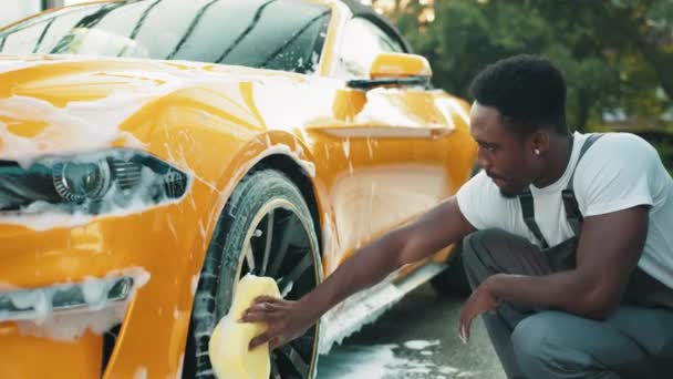 Voiture sport moderne de luxe en mousse de savon à l'extérieur au service de lavage de voiture. Vue latérale d'un beau jeune Africain utilisant une éponge jaune pour nettoyer les jantes à l'extérieur. Concept de voiture propre. Moussant après frottement — Video
