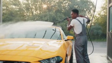 Tişörtlü ve koruyucu tulumlu yakışıklı Afrikalı adam yüksek basınçlı su pompasıyla sarı lüks spor arabasını yıkıyor. Elle oto yıkama, basınçlı su, dışarıda araba yıkama. Araba yıkama .