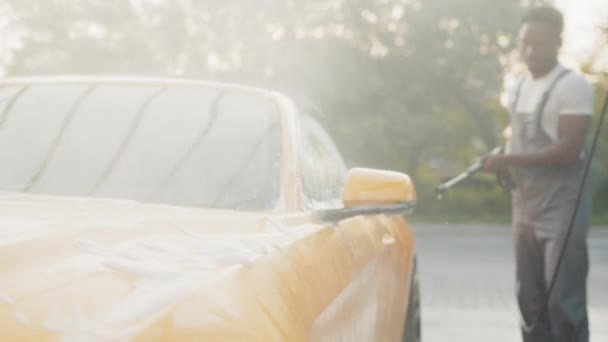 Przystojny Afrykanin w t-shircie i szarym kombinezonie myjący swój żółty luksusowy samochód, płukający mydło strumieniem wody pod wysokim ciśnieniem. Ręczna myjnia samochodowa z wodą pod ciśnieniem w myjni samochodowej na zewnątrz. — Wideo stockowe