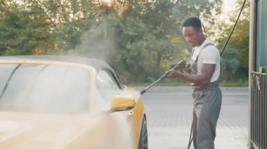 Dışarıdaki oto yıkama servisinde yüksek basınçlı suyla dikiz aynası yıkıyorum. Dikiz aynası dışında su jeti ile yıkanan odaklanmış Afrikalı bir adam. Arkadaki adama odaklan. Araba yıkama.