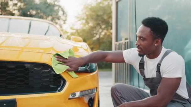 Porträtt av en leende ung afroamerikansk man i grå overaller och vit t-shirt, rengöring modern sportbil huva med gröna mikrofiber tyg utomhus, vid självbilstvätt station — Stockvideo