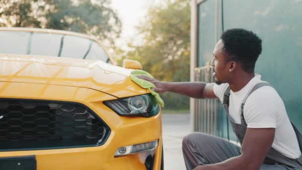Porträtt av en leende ung afroamerikansk man i grå overaller och vit t-shirt, rengöring modern sportbil huva med gröna mikrofiber tyg utomhus, vid självbilstvätt station — Stockvideo