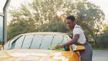 Açık hava self servis istasyonunda oto yıkama ve temizlik. Yakışıklı, sakallı, genç Afrikalı bir işçi yaz sabahı açık havada yeşil bir mikrofiber bezle sarı araba kaputunu temizliyor..