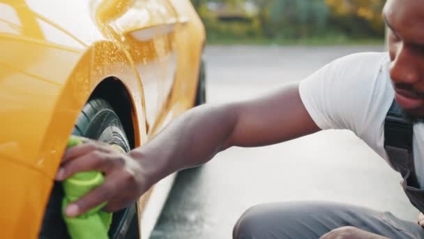 Autowaschanlage im Freien. Selbstbedienungskonzept für Autowäschen. Schöner afrikanischer Typ, der während des Waschvorgangs im Freien das Rad eines modernen Luxusautos mit grünem Mikrofasertuch wischt. Konzept für saubere Autos — Stockvideo