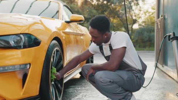 Young worker of self car wash service African bearded man cleaning a wheel, car rims of modern luxury yellow sport car with green microfiber cloth, at outdoor car wash self service. Clean car concept — Stock Video