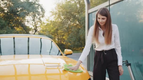 Porträtt av en leende ung kaukasisk affärskvinna på vit skjorta och svart byxa, rengöring modern sportbil huva med gröna mikrofiber tyg utomhus, vid självbilstvätt station. Begreppet ren bil — Stockvideo