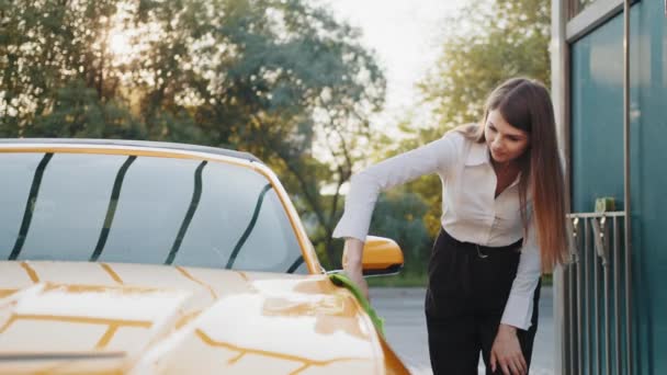 屋外のセルフサービスステーションでの車の洗浄とクリーニング。成功したビジネスのショット女性は夏の晴れた朝に屋外の緑のマイクロファイバーの布で彼女の黄色の車のフードを掃除します。クリーンカーコンセプト — ストック動画