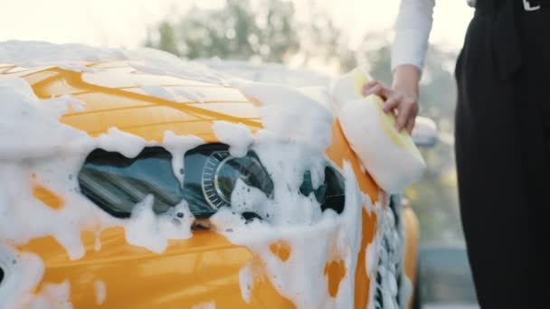 Närbild beskärs bilden av handen av unga kaukasiska kvinna med gul svamp tvätthuva och strålkastare av hennes bil på en självbetjäning biltvätt utomhus. Lyxbil täckt av skum. Biltvätt utomhus. — Stockvideo