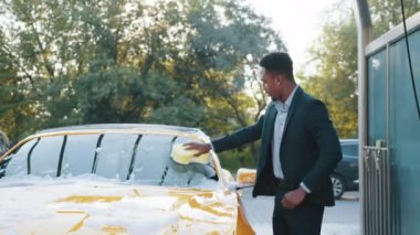 Yakışıklı, Afro-Amerikan iş adamı. Araba yıkama servisinde arabasını sarı sünger ve sabun köpüğüyle temizliyor. Temiz araba konsepti. Dışarıda araba yıkama. Sürttükten sonra köpürme