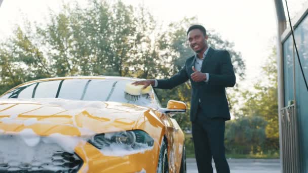 Guapo joven empresario afroamericano limpiando su parabrisas de coche con esponja amarilla y espuma de jabón al aire libre en el servicio de lavado de coches y mostrando el pulgar hacia arriba. Lavado de autos al aire libre. Espuma frotando el coche. — Vídeos de Stock