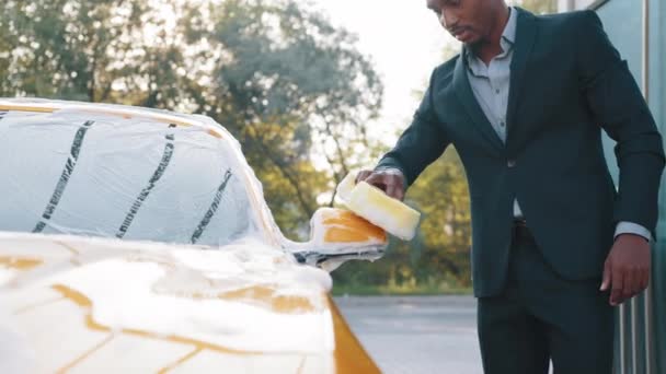 Çekici Afrikalı Amerikalı genç işadamı, modern lüks arabasının dikiz aynasını temizleme köpüğü ve sarı süngerle açık hava oto yıkama istasyonunda yıkıyor. — Stok video