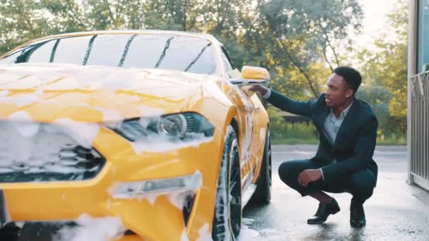 Lyxig gul bil i tvålskum utomhus vid biltvättservice. Sidovy av stilig ung afrikansk affärsman som använder gul svamp för rengöring bildörr med tvålskum. Skum efter gnidning av bilen. — Stockvideo