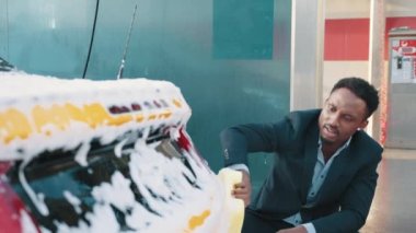 Açık havada oto yıkama servisinin yatay görüntüsü. Yakışıklı Afro-Amerikalı takım elbiseli adam arka farları sarı sünger ve köpükle yıkıyor.