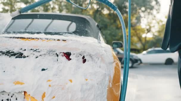 Машина покрыта пеной. Автомобиль моется мылом на автомойке. Автомойка с моющим средством высокого давления. Мыть на открытом воздухе концепции. Желтая машина в пене, моется с мылом — стоковое видео