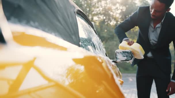 Joven atractivo hombre afroamericano en traje de negocios lavado Espejo exterior retrovisor de su moderno coche de lujo en una estación de lavado de automóviles de autoservicio al aire libre con espuma de limpieza y esponja amarilla — Vídeos de Stock
