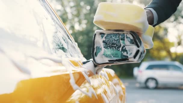 Närbild beskärda bilden av handen av ung afrikansk affärsman med gul svamp tvätt backspegel av sin bil på en självbetjäning biltvätt utomhus. Lyxbil täckt av skum — Stockvideo