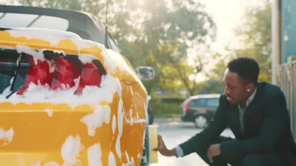 Luxus modern sport autó szappanhab szabadban autómosó szolgáltatás. Oldalnézet jóképű fiatal afrikai üzletember használja sárga szivacs tisztítására felnik szabadban. Habzás a kocsi dörzsölése után — Stock videók