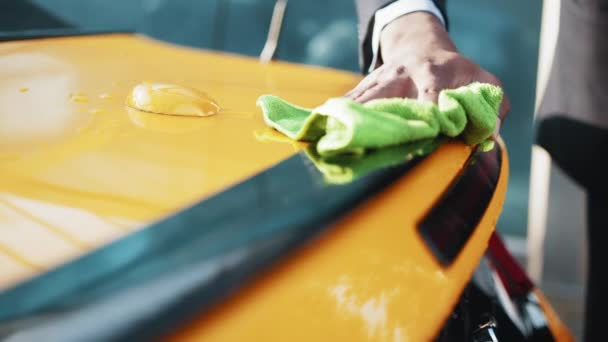 Un homme essuie l'humidité et les taches de sa voiture avec un chiffon spécial. Les mains en gros plan. Homme d'affaires décontracté en tenue formelle essuyant le dos de sa voiture jaune avec un tissu en microfibre verte au lavage de voiture en plein air — Video