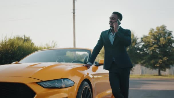 Homem africano sério em terno de negócios segurando smartphone moderno na mão enquanto está perto de luxo carro cabriolet amarelo. Jovem macho em terno de negócios posando ao ar livre e olhando para o lado — Vídeo de Stock