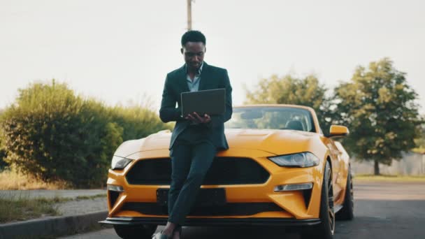 Schöner afroamerikanischer Mann im Business-Anzug. Geschäftsmann arbeitet mit Laptop. Mann steht neben seinem gelben Sportwagen auf der Grünfläche. — Stockvideo