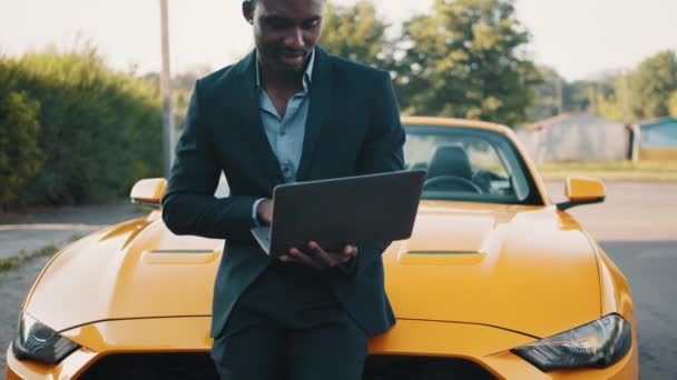 Schöner junger afroamerikanischer Geschäftsmann, der mit Laptop arbeitet, während er in der Nähe eines modernen gelben Cabriolets im Freien steht, schöner afrikanischer Mann auf dem Hintergrund eines Sportwagens. — Stockvideo