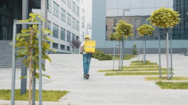 Genç kurye e-scooterına biniyor. Erkek paket teslimatçısı sokağın aşağısındaki sarı sırt çantalı boş kaldırımdan elektrikli scooter kullanıyor. Mal teslimatı.