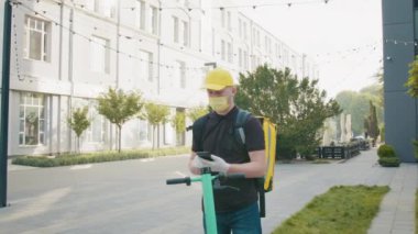 Karantina sırasında teslimat. Bir apartmanın arka planında akıllı telefonu olan bir teslimatçı ya da modern ofis, tıbbi eldiven ve maske takmış. Elektrikli scooter kullanan kurye..