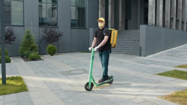 Entrega hombre con mochila amarilla y máscara protectora montar scooter eléctrico a través de la ciudad con entrega de alimentos, fondo del centro de negocios. Excelente entrega durante la cuarentena. — Vídeos de Stock