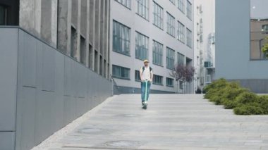 Gözlüklü genç bir adam, teslimat servisinde çalışıyor, sarı sırt çantası ve beyzbol şapkası var, elektrikli scooter kullanıyor. Teslimatçı şehirde yiyecek getiren bir scooter kullanıyor.