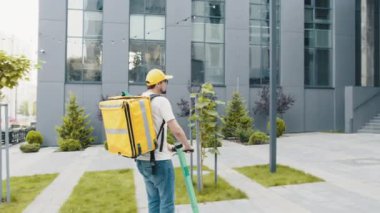 Tanımlanamayan genç kurye e-scooterıyla geziyor. Erkek paket teslimatçısı, elektrikli scooter ile sokakta sarı sırt çantasıyla geziyor. Ekolojik nakliye aracı.