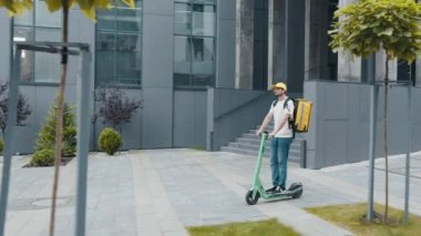 Termal çantalı ya da sırt çantalı sakallı teslimatçı şehirde elektrikli scooter 'a binip yiyecek getiriyor. Kurye, teslimat hizmeti konsepti. Yan görünüm, mal teslimatı ekolojik taşımacılık
